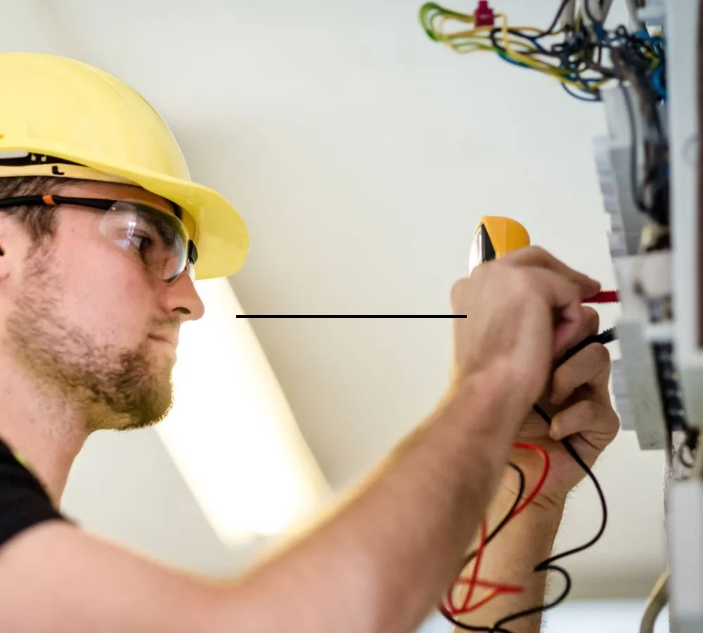 An electrician in a hard hat and safety glasses testing at a distribution board with a clamp meter