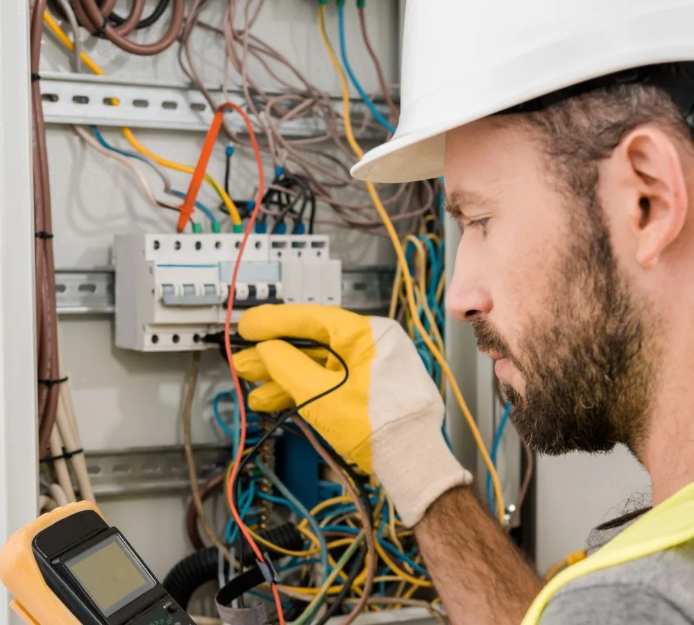 A technician in a hard hat and work gloves using a multimeter inside an open switchgear panel