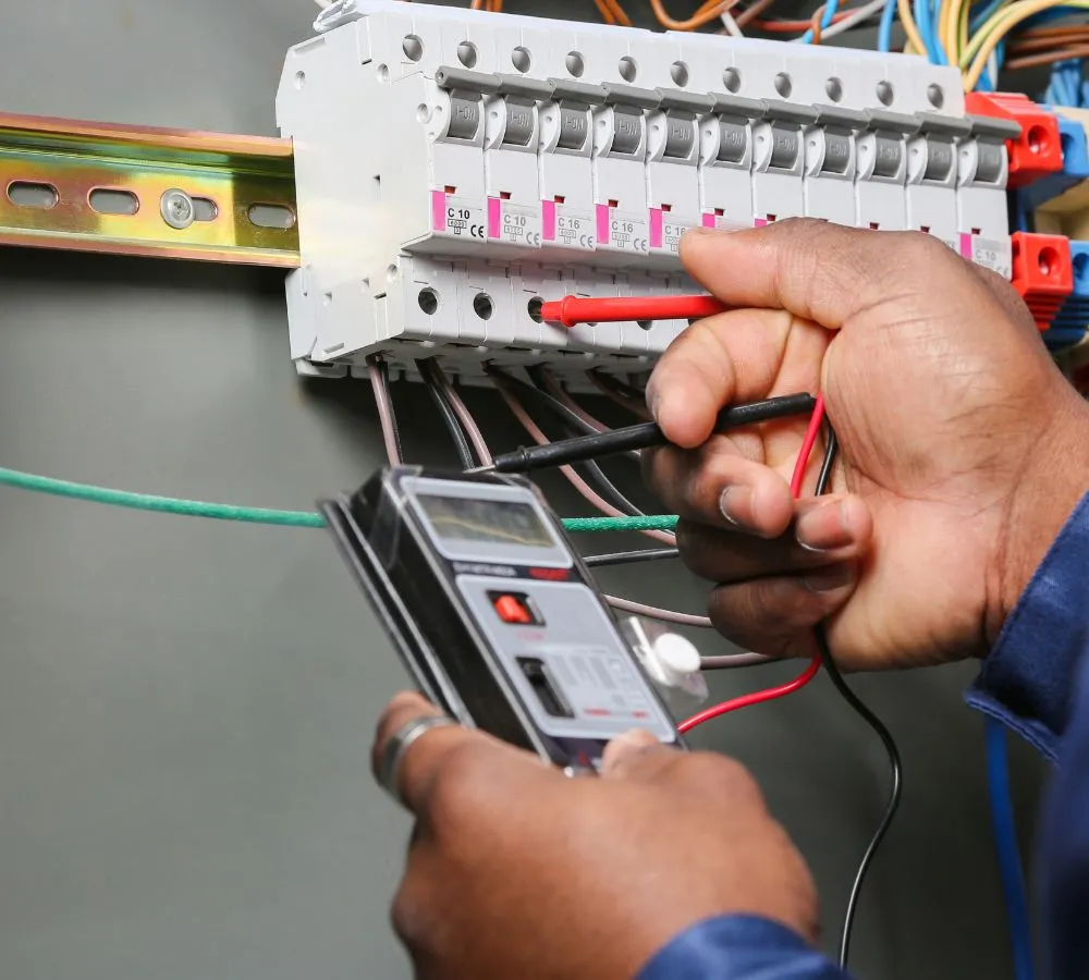 An electrician taking a reading at a distribution board, test leads on a DIN rail of C10 and C16 circuit breakers