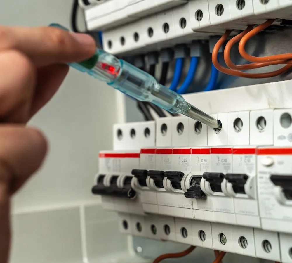 A voltage tester held against the terminals of a consumer unit, with rows of miniature circuit breakers and RCBOs below